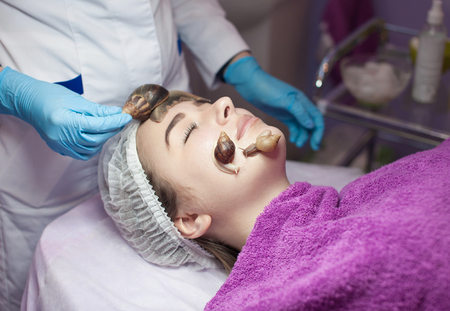 Young woman receiving snail facial massage. Snail on face. Cleaning procedure in spa salon. Blurred background.の写真素材