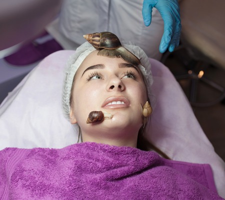Young woman receiving snail facial massage. Snail on face. Cleaning procedure in spa salon. Blurred background.の写真素材