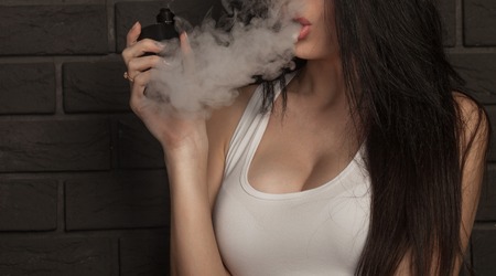 Young beautiful brunette woman smoking, vaping e-cigarette with smoke on black background.の写真素材