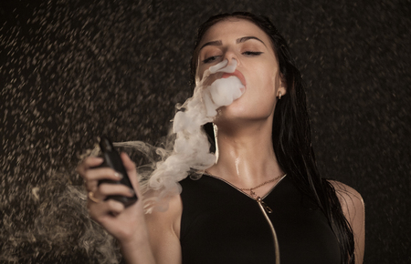 Young beautiful woman vaping e-cigarette. Water flowing on woman face.の写真素材