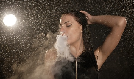 Young beautiful woman vaping e-cigarette. Water flowing on woman face.の写真素材