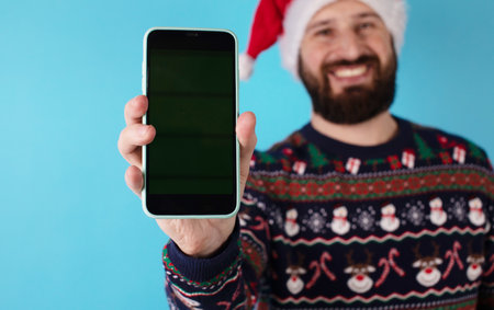 Young bearded man in a Santa Claus hat holds a smartphone with a green screen on a blue backgroundの写真素材