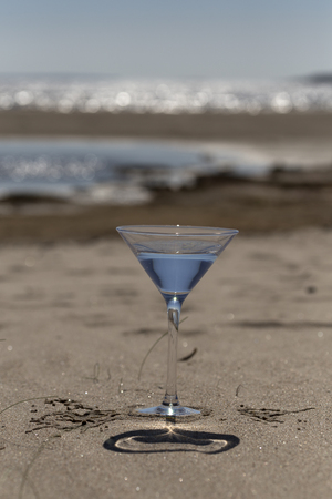 A glass of water on a sandy beach with parts of the ocean in the backgroundの写真素材