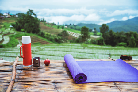 Purple yoga mat and red water bottle with a glass of water stainless Put forward. And Red apple on wooden background. On the balcony landscape Natural Field.Travel relax. papongpieng in Thailandの写真素材