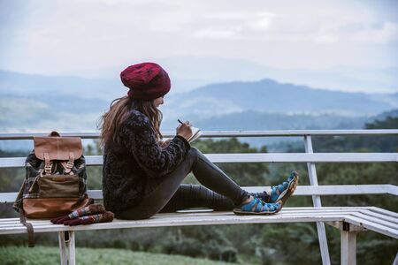 The girl is sitting relaxing at the natural mountain viewpoint and writing a record of her winter travel. smiled happy writing a diary.の写真素材