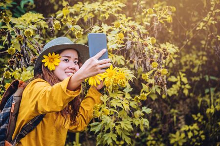 A girl with a backpack is using the phone to take selfie a picture of the Bua Tong flower yellow. "Mexican sunflower"の写真素材