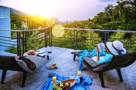 A happy woman sitting reading relaxing book on the balcony at home, summer on holiday drinking wine,orange juice and fruit on the terrace in the at sunset.の写真素材