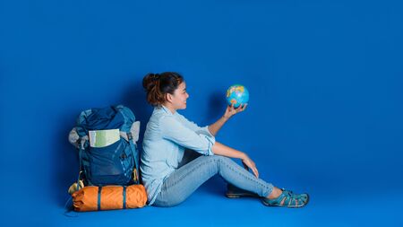 Tourist planning vacation with the help of world map with other travel accessories around. Woman traveler with suitcase on Blue color background. Girl with a ball globe in the hand. Travel backpackの写真素材