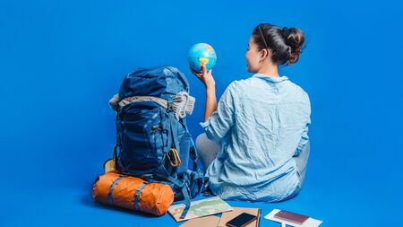 Tourist planning vacation with the help of world map with other travel accessories around. Woman traveler with suitcase on Blue color background. Girl with a ball globe in the hand. Travel backpackの写真素材