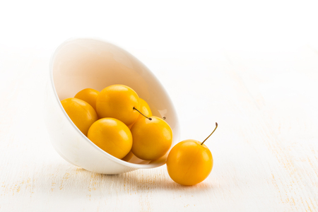 Handful of yellow cherry plums in ceramic bowl on white wooden surfaceの写真素材