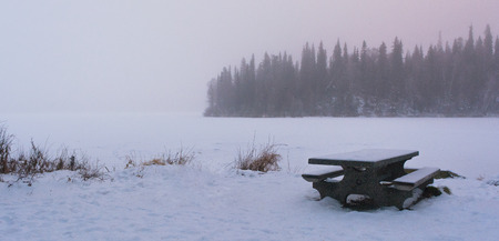 Snow covers a park bench on a snow covered frozen lake.