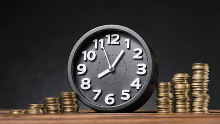round clock between the increasing coins on wooden desk against black backdrop. Resolution and high...