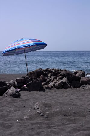 Overlooking a quiet beach where in the foreground you can see an umbrella and some rocks, in the background the blue sea and skyの写真素材