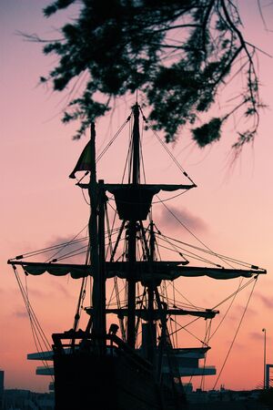 Silhouette image of a 15th century Spanish caravel boat framed by tree branches while moored in the...