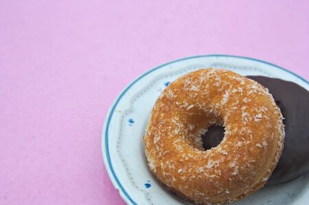 Two doughnuts one with coconut and one with chocolate on top on a plate and a pink background to add...