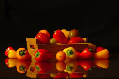 A few small peppers of red and yellow colors in a small wooden box that is on a table where the peppers are reflected. Dark background, dark foodの写真素材