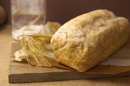 A loaf of homemade whole wheat bread next to a wheat branch and on a wooden board