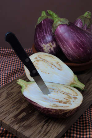 A still life of aubergines, on a wooden board, in which one of them is split in half and has a small knife stuck in its centerの写真素材