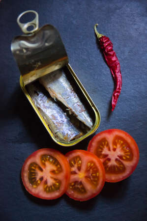 Closeup of a can of sardines next to some red tomatoes and a chilli on a black slateの写真素材