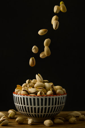 A bunch of pistachios falling on a bowl full of pistachios on a wooden table and dark backgroundの写真素材