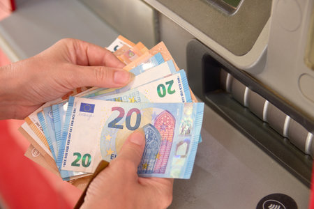 The hands of a woman hold several euro bills in front of an ATM in a bankのeditorial素材