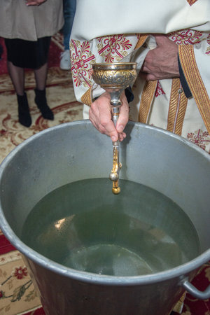 an orthodox priest inserts a cross into the metal baptismal vessel in Romania