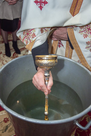 an orthodox priest inserts a cross into the metal baptismal vessel in Romania
