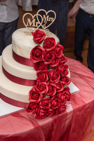 Three-tiered wedding cake decorated with red roses and white frosting, each tier is surrounded by a red satin ribbon.
At the top is a wooden heart ornament with the words "Mr & Mrsの写真素材