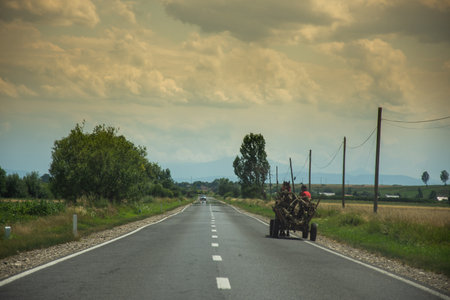 A horse-drawn cart moves along a paved road, somewhere in Romania. In the background, a landscape of hills and vegetation is visible, under a cloudy sky.の写真素材