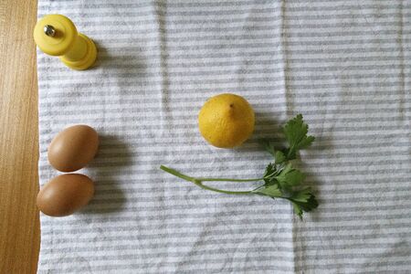 Cenital shot of 2 eggs, parsley, pepper shaker and a lemon over a kitchen towel on a wooden tableの写真素材