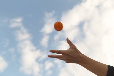 Front Shot of young hand throwing an organic tangerine in the air on a blue sky with clouds. Copy Spaceの写真素材