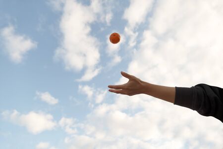 Front Shot of a young hand throwing an organic tangerine in the air on a blue sky with clouds. Copy Spaceの写真素材