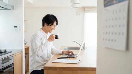 Young asian man using laptop computer and drinking coffee in kitchen.の素材