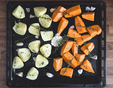Fresh raw potatoes, sweet potatoes and garlic with some herbs on a black oven tray. Photograph taken from above.の写真素材
