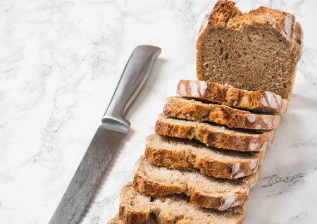 Brown loaf of bread sliced with a silver knife on the side. Marble counter. Angle view of the slices