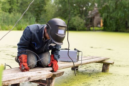 man welder in blue uniform works, welding mask and protective red gauntlets, metal welding with a...