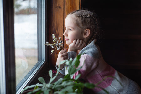 Beautiful girl looks out the window with a smile. Caucasian little girl with willow branches. Stay indoors. The arrival of spring. Mother's day.の写真素材