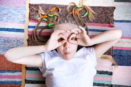 A Caucasian girl with pigtails, lies on a floor on a woven striped rug, shows glasses from folded...