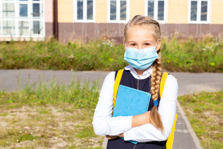 A girl in a blue school uniform stands in front of school building in a medical mask, holds a...