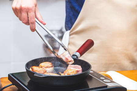 Meat in bacon is fried in a cast-iron frying pan, a man chef in an apron turns the meat over with metal tongs.の写真素材