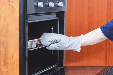 A hand in a protective glove pulls out a cooked hot meat dish from the oven. Safety concept. Kitchen, food preparationの写真素材