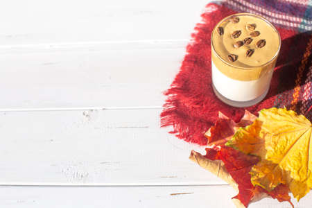 Traditional Korean caffeinated milk drink, Dalgona coffee in a glass with coffee beans on a white board background. Dry maple leaves. Warm checkered scarf or plaid. Autumn breakfast in cafe.Copy spaceの写真素材