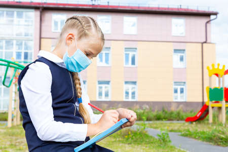 A girl in a blue school uniform and a medical mask sits and writes with a pen in a notebook outdoors against background of school building and playground. New school year, new normal. Copy spaceの写真素材