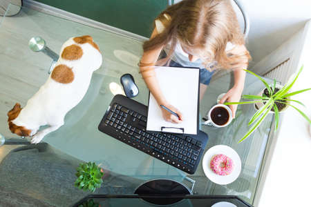 Girl sits at a glass table with a computer, holds a pen over a blank white of paper, a dog lies nearby, donut and coffee. Home office, distance learning, remote work. Breakfast at workplace. Top viewの写真素材