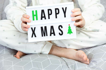 A child in white pajamas with asterisks sits cross-legged on the bed and holds a light board with the inscription happy xmas and Christmas tree in his hands. Cozy home, bedroom. Christmas morning.の写真素材