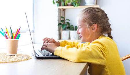 Caucasian blonde school girl, typing on a laptop keyboard while sitting by the window at home,...