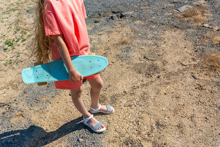 A slender teenage girl with a skateboard in her hand in a summer trendy suit with fluttering blonde hair against a background of cracked ground on a sunny summer day. Healthy lifestyle. Copy space.の写真素材