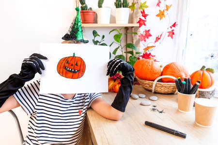 A child shows his drawing for Halloween, sitting at a desk by the window, surrounded by pumpkins and houseplants. Sketch drawing of an orange laughing pumpkin with sharp teeth. Get ready for the partyの写真素材