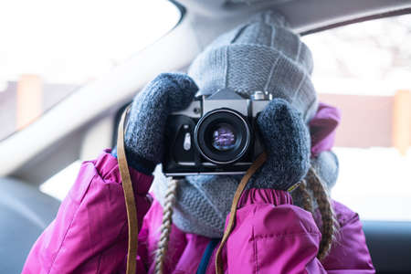 A girl in winter clothes takes a picture with a camera while in the car. Road travel in winter by car. Vintage film SLR camera on a leather strap in hands. Portrait lens.の写真素材