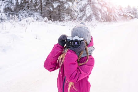 Backlit portrait of girl in warm clothes taking photo in winter forest. Child with camera for walk in winter. Day of photographer. Hobby, photography school. Lens looking at camera.の写真素材